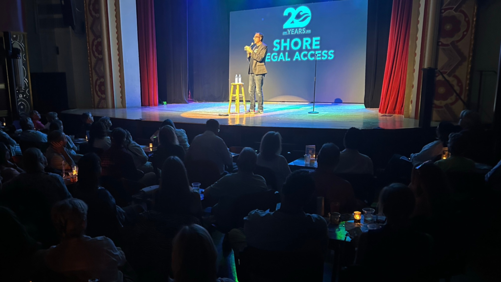 A man speaks into a microphone on a stage in front of a screen reading 20 Years Shore Legal Access, viewed by an audience in a darkened theater.
